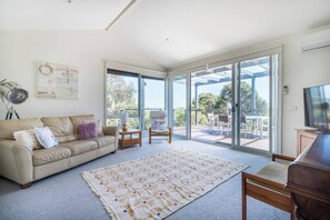 A spacious, air-conditioned living area upstairs is connected to the first bedroom, featuring a comfortable sofa, piano, and access to the deck for ocean breezes.
