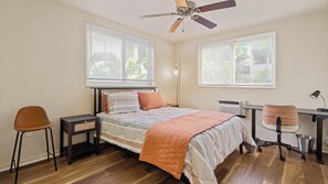 Primary bedroom. Queen size bed with linens provided. Window AC unit and ceiling fan. Standing desk to accommodate remote workers. Nightstands, floor lamp, and chair.