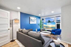 Relax in the cozy living room featuring a modern blue accent wall, plush seating, and a large flat-screen TV with natural light pouring in.