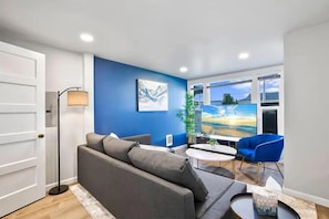 Relax in the cozy living room featuring a modern blue accent wall, plush seating, and a large flat-screen TV with natural light pouring in.