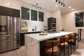  A modern kitchen with a large island, a stainless steel refrigerator, and dark cabinets. The kitchen has a large window with a view of the city. 