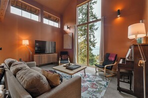 The living room boasts a stunning 14-foot-tall scenic window with views of the water and mountains, along with high ceilings, a smart TV, a piano, and a spacious coffee table perfect for games, drinks, and snacks.