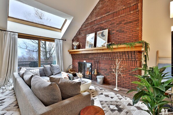 Cozy living room of the Sunrise Timberline ski-in ski-out rental condo in Killington, VT, featuring a large L-shaped sofa, a balcony and a high window showing mountain views, and an exposed-brick wall with a fireplace.