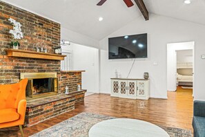 Living room with brick fireplace, orange chair, mounted TV, white console…