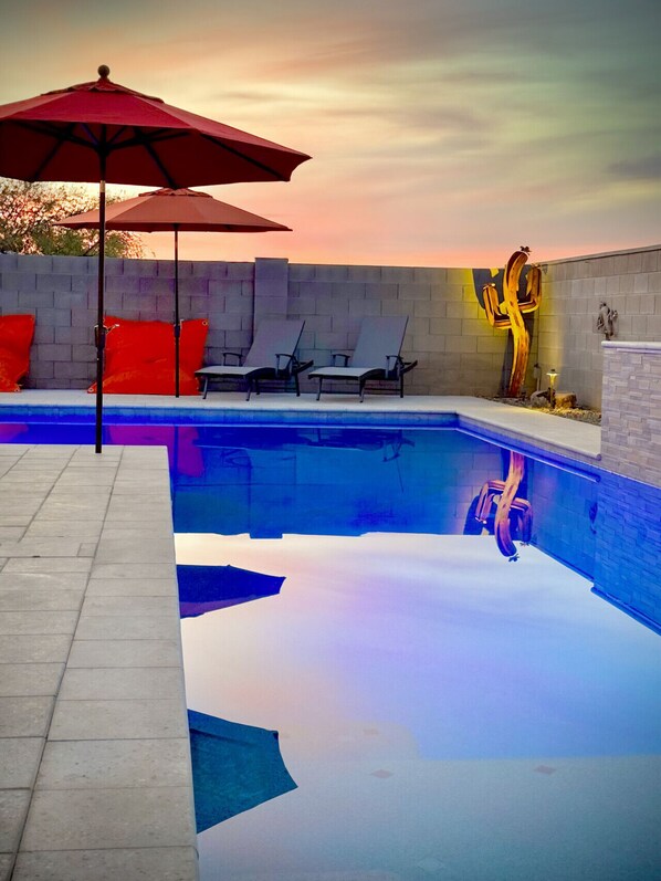 A shimmering blue pool reflects a golden sunset, with relaxing lounge chairs.