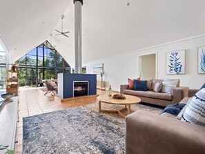 A living room with a high, vaulted ceiling and large windows that let in ample natural light. Furnished with two large, comfortable-looking sofas arranged around a coffee table, and a fireplace with blue tile is the centerpiece of the room.