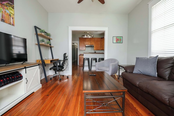 View from the front of the house, showing the living room with a couch that spans the entire room, small workspace, and part of the full kitchen in the next room.