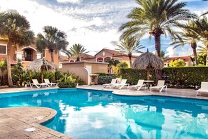 A beautiful tropical pool area surrounded by lush greenery and tall palm trees. The crystal-clear water reflects the blue sky above, while lounge chairs and thatched umbrellas create a relaxing atmosphere. In the background, colorful villas with terracott