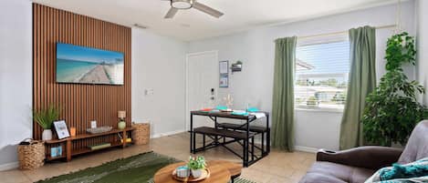 Bright and breezy with tropical charm, this cozy living space features a smart TV, comfy seating, modern accents, and a dining nook by a sunlit window—perfect for relaxing, dining, or enjoying your favorite shows in comfort and style.