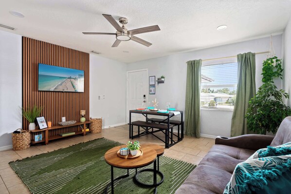 Bright and breezy with tropical charm, this cozy living space features a smart TV, comfy seating, modern accents, and a dining nook by a sunlit window—perfect for relaxing, dining, or enjoying your favorite shows in comfort and style.