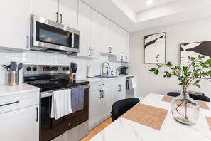 Modern kitchen featuring sleek white cabinets and stainless steel appliances, including an oven, microwave, and countertop. The kitchen opens into a dining area with a stylish white marble table and four chairs, providing a comfortable space for guests to
