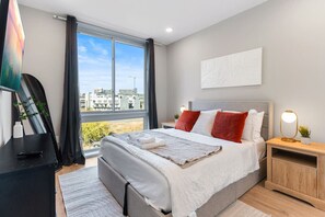 A modern bedroom with a queen-sized bed and a large window with darkening shades, offering views of the city skyline.