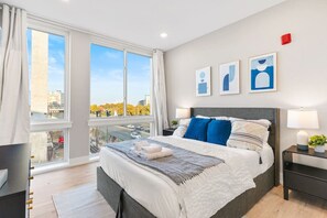 A modern bedroom with a queen-sized bed and a large window with darkening shades, offering views of the city skyline.