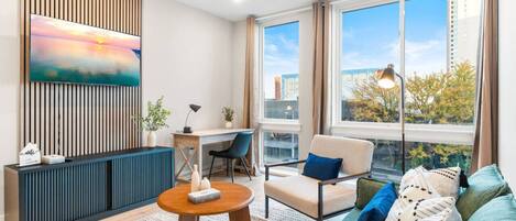 A cozy living room featuring a plush couch, modern coffee table, dedicated workspace, TV, and large windows that bathe the room in natural light.