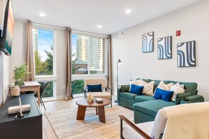 A cozy living room featuring a plush couch, modern coffee table, dedicated workspace, TV, and large windows that bathe the room in natural light.