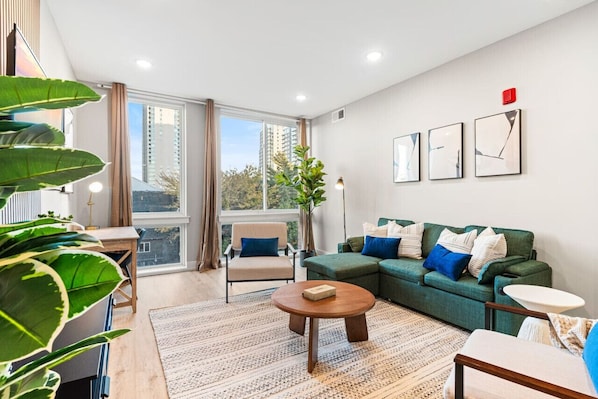A cozy living room featuring a plush couch, modern coffee table, dedicated workspace, TV, and large windows that bathe the room in natural light.