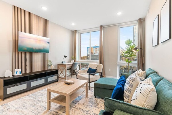 A cozy living room featuring a plush couch, modern coffee table, dedicated workspace, TV, and large windows that bathe the room in natural light.