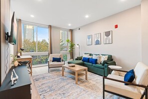 A cozy living room featuring a plush couch, modern coffee table, dedicated workspace, TV, and large windows that bathe the room in natural light.