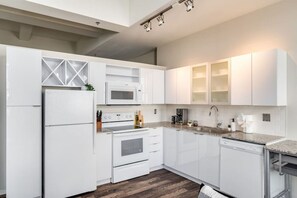 This modern kitchen features a spacious island with cabinetry, sleek white countertops, stainless steel appliances, open shelving, and stylish lighting.