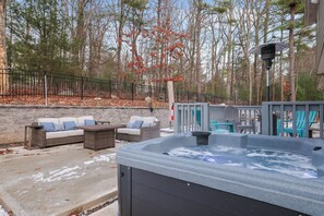 Relaxing outdoor space featuring a hot tub, cozy patio seating with cushioned furniture, and a firepit, all set against a tranquil wooded backdrop. Perfect for unwinding year-round.