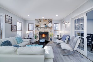 Cozy living room with a stone fireplace, white sofas, modern chairs, and pops of blue décor. Bright, stylish, and perfect for relaxing or entertaining.