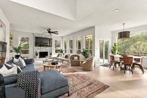 Elegant living room with a cozy gray sectional, abstract wall art, a rustic stone fireplace with a mounted TV, stylish accent chairs, a patterned area rug, built-in shelving, and large windows flooding the space with natural light.