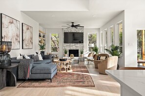 Elegant living room with a cozy gray sectional, abstract wall art, a rustic stone fireplace with a mounted TV, stylish accent chairs, a patterned area rug, built-in shelving, and large windows flooding the space with natural light.