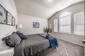 This fourth bedroom boasts a comfy queen-sized bed. Large windows with blinds let in natural light, complemented by a side lamp and half-body mirror. Framed artwork adorns the wall, and carpeting adds warmth and coziness to the floor.
