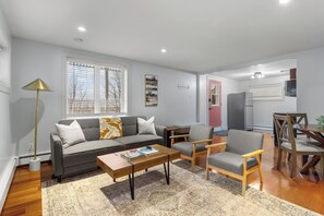 Living space with a comfortable gray sofa, two stylish armchairs, and a wooden coffee table. The room features a large window with blinds, a unique floor lamp, and tasteful wall art. The open layout leads to a dining area and a kitchen with a stainless steel refrigerator and a red door, creating a welcoming and functional environment. Perfect for relaxing and enjoying your stay.