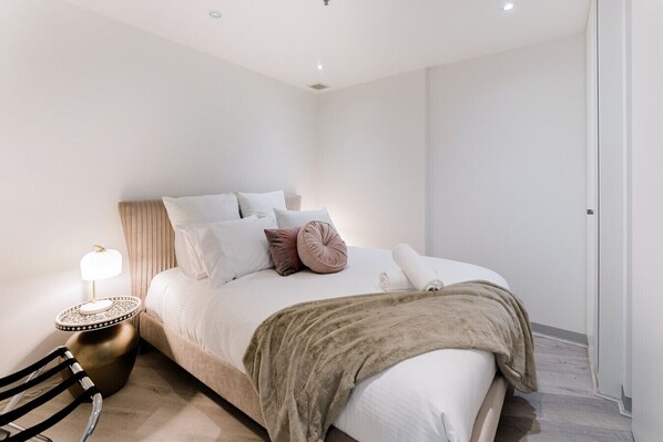 Cozy bedroom with a plush neutral-tone bed, soft pillows, and a warm blanket, complemented by a chic bedside lamp and modern decor.