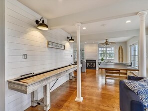 This luxurious beach house features an inviting game room with a shuffleboard table, perfect for entertaining while enjoying the elegant shiplap decor and natural light from expansive windows.