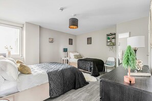 Bright and modern bedroom featuring two beds with black and white bedding, hardwood floors, natural light from large windows, and minimalist decor including small plants.