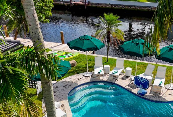 Elevated view of the sparkling pool, lush greenery, and canal, framed by palms.