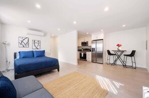 queen-sized bed dressed in rich blue bedding, featuring modern art above the bed and a sleek floor lamp for added ambiance. The room's open layout flows seamlessly into the living area, making it both spacious and inviting.