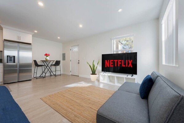 A cozy living area with a gray sofa bed, large TV ready for Netflix, a small dining nook, and a stainless steel fridge.