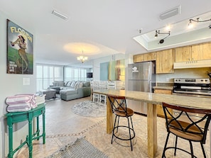 Stylish kitchen with breakfast bar flows into a spacious dining and living area, featuring coastal décor and ample natural light for a welcoming and functional layout.