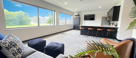 A spacious lounge area bathed in natural light from wall-to-wall windows, featuring a cozy sectional, stylish poufs, and modern seating. Perfect for relaxing or entertaining with scenic views and seamless access to the kitchen and bar.
