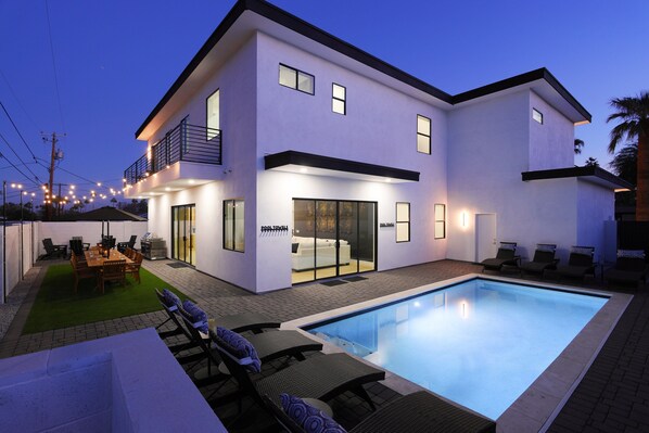 Resort-style backyard with a sparkling pool, sun loungers, an outdoor dining table for 12, a grill, and a cozy fire pit with surrounding seating.