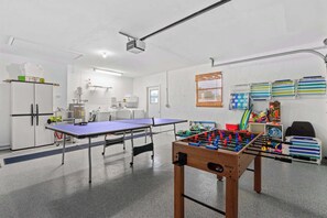 Spacious multi-purpose room featuring a ping pong table and a foosball table, perfect for indoor entertainment. The floor has a smooth finish, and a large refrigerator is visible in the background alongside other storage item