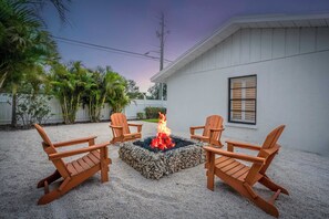Captured at dusk, this image highlights the fire pit in full glow, casting warm light around the seating area. The chairs are perfectly positioned for gathering, while the soft twilight sky 
