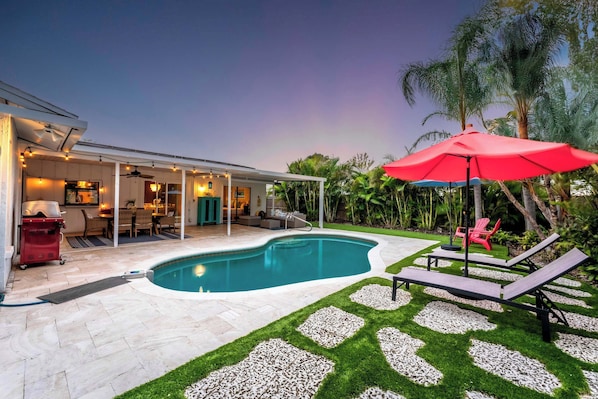 Captured at dusk, this photo reveals the enchanting ambiance of the pool area with warm lighting illuminating the space. The red umbrella and cozy seating area invite relaxation as the sun sets, creating a perfect evening retreat.