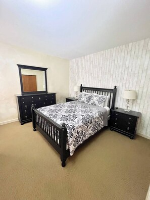 This cozy bedroom features a classic black wooden bed frame with matching dresser and nightstands. A delicate floral-patterned bedspread complements the soft, neutral tones of the room for a welcoming and restful retreat.