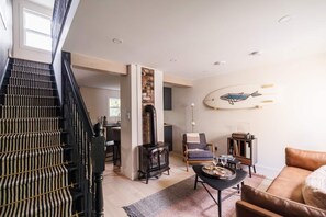 This living space blends modern and rustic charm. A  leather couch, coffee table, and vintage wood stove create a warm, inviting vibe. The surfboard wall art adds a playful touch, while the staircase enhances the space’s character #QVTides







