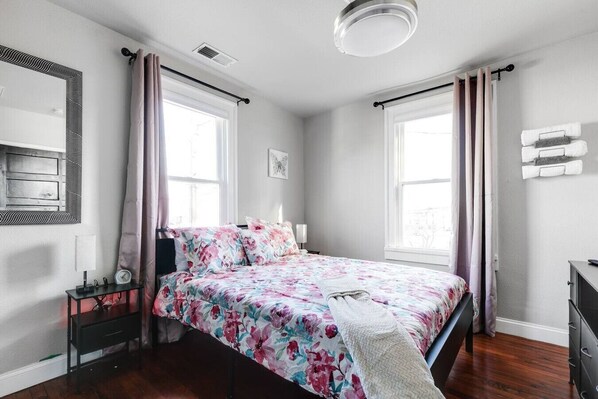 Queen comfort and soft floral aesthetics create a serene retreat with two large windows, a nightstand, a Roku TV, a large mirror, and a chest of drawers. A spacious hanging rack offers additional storage.