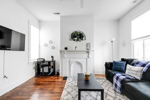 A fresh and inviting space with large windows for natural light, a cozy couch, a coffee table, and a flat-screen Roku TV. Includes a dedicated work nook with a fan and board games. Conveniently located across the hall from the kitchen.