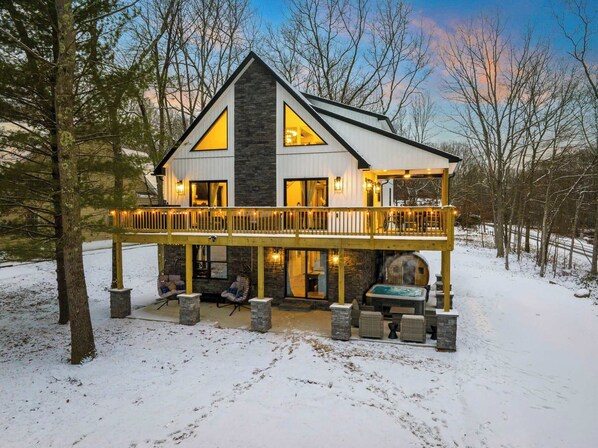 A modern mountain retreat stands gracefully against a snowy backdrop, with tall windows and warm glow lighting up the wraparound deck.