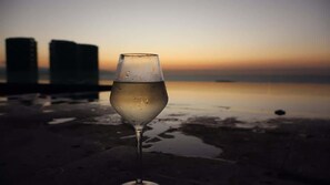 A chilled glass of white wine, droplets glistening, rests by an infinity pool reflecting a golden sunset fading to dusky purple. Modern towers and the calm ocean complete the serene, luxurious scene.