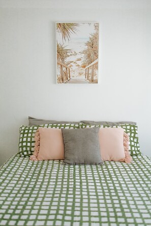 A cozy bedroom with a green and white checkered bedspread, decorative pillows, and a framed coastal scene hanging on the wall.
