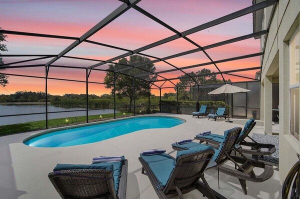 Beautiful sunny pool deck with a covered lanai and dining area, sun loungers, and a serene lake view.