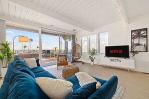 A stylish living room designed for comfort and leisure, featuring a plush deep blue velvet sofa, which is complemented by soft throw pillows and a chic floor lamp. A sleek white entertainment unit with a large flat-screen TV displaying Netflix ensures gue